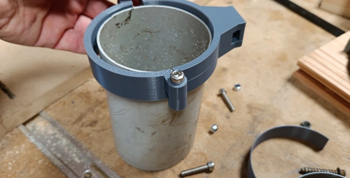 Sur l'établi : le collier posé autour d'une chute de descente, son logement carré visible en haut, la visserie inox et un second collier autour