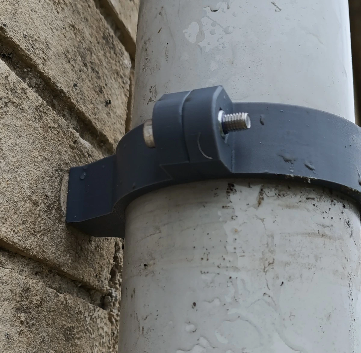 Vue de trois quarts : la patte du collier vient en appui contre le mur de pierre, et la vis inox traverse les deux oreilles